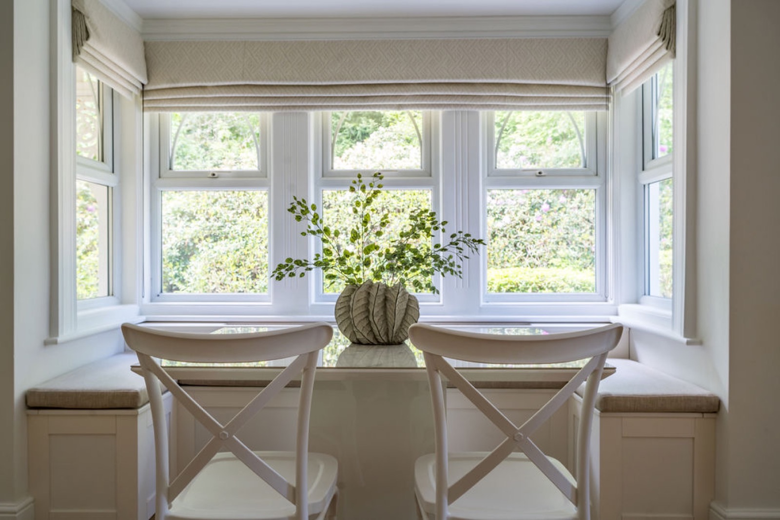 An intimate breakfast nook set into a bay window, with a bespoke banquette, cross-back chairs and a sculptural vase — a considered corner that makes the everyday feel special.